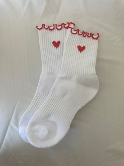 A pair of white crew-length socks with a red heart jacquard design and lettuce-edge trim.